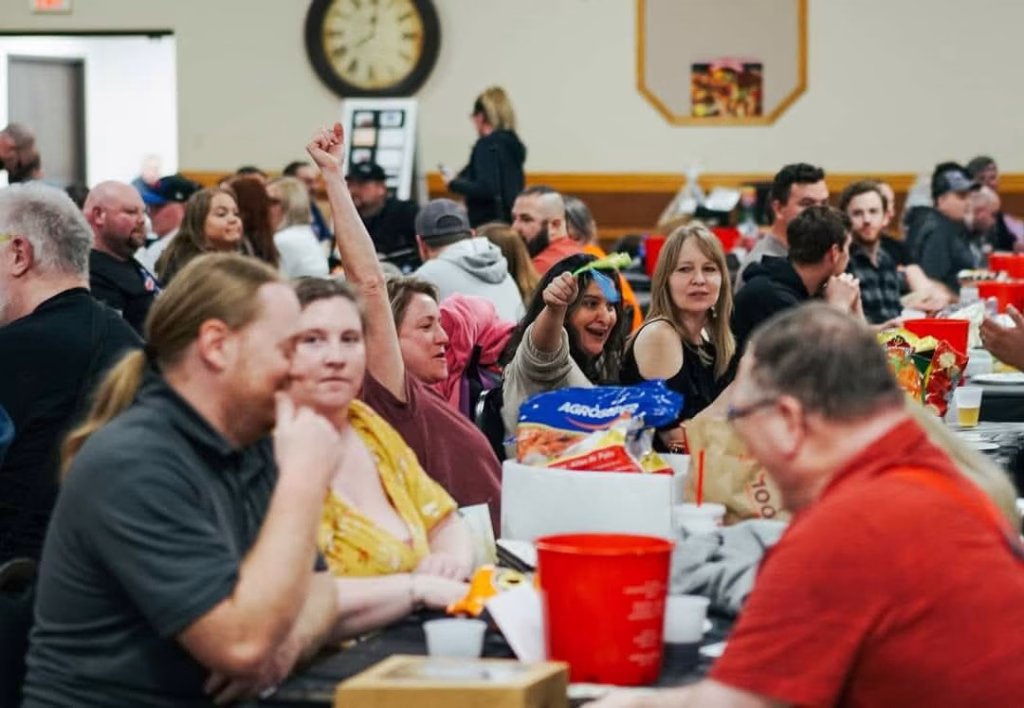 A large group of people sit at tables in a community hall. One woman in the middle raises her arm enthusiastically and smiles, while others around her chat and eat. There are snacks and drinks on the tables.