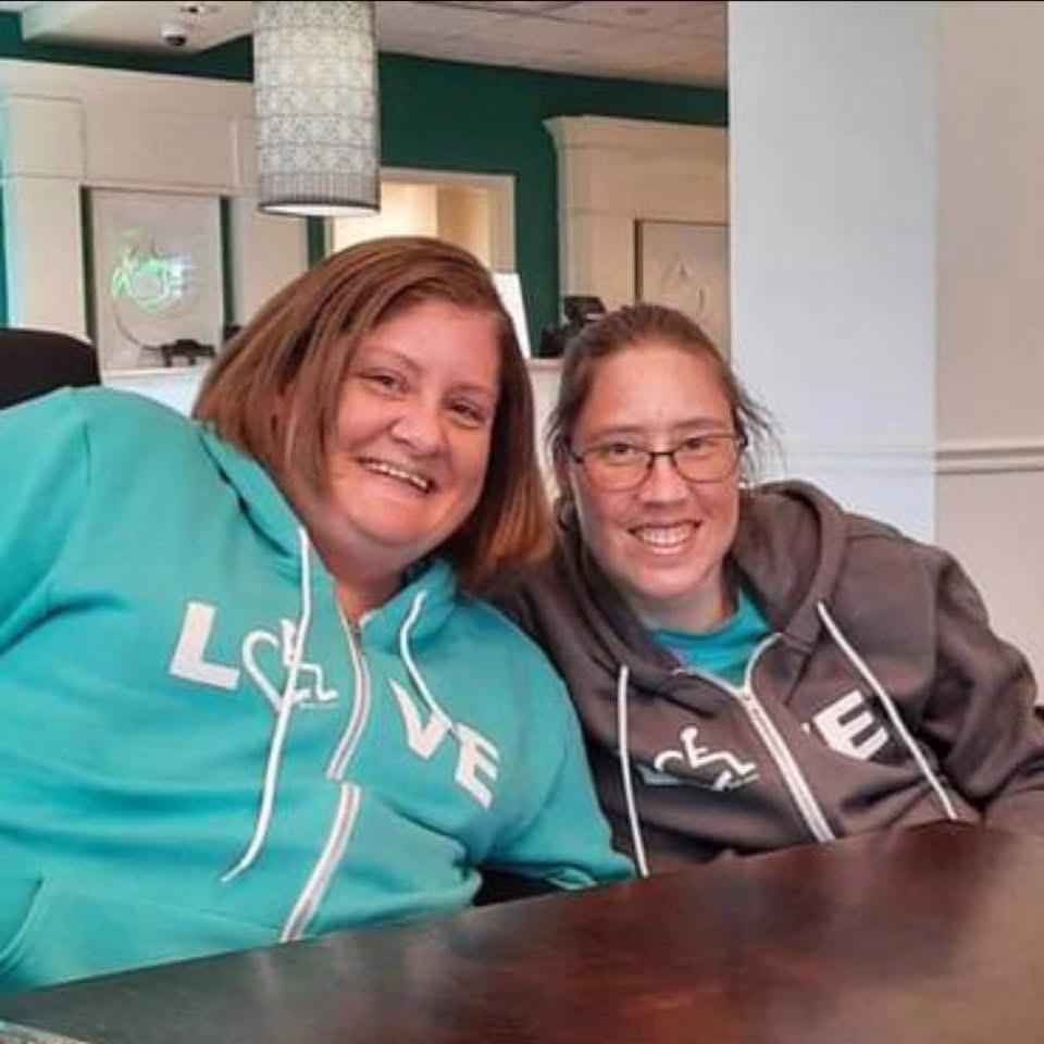 Two women sit side by side at a table, smiling at the camera. Both are wearing teal and gray hoodies that spell LOVE with a wheelchair symbol as the “O”, in a brightly-lit room with teal and white walls.